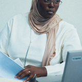 A woman wearing a beige hijab and glasses, dressed in a white top, sitting at a desk with a laptop and holding papers.
