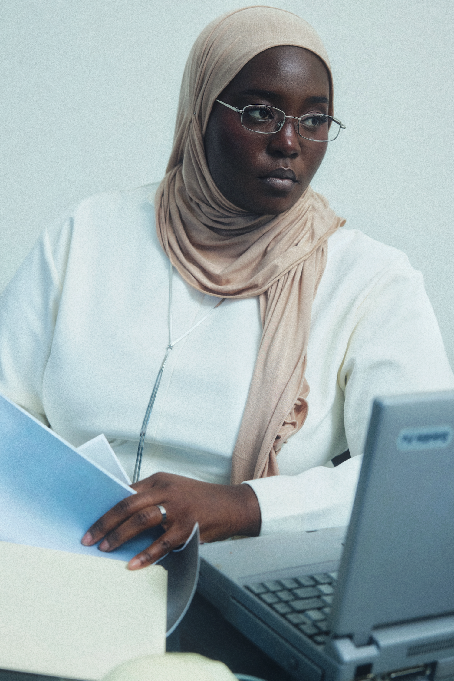 A woman wearing a beige hijab and glasses, dressed in a white top, sitting at a desk with a laptop and holding papers.