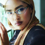 A close-up lifestyle shot of a woman wearing a beige and brown patterned hijab, silver-framed eyeglasses, and a black leather jacket, resting her head on a reflective surface.