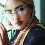 A close-up lifestyle shot of a woman wearing a beige and brown patterned hijab, silver-framed eyeglasses, and a black leather jacket, resting her head on a reflective surface.
