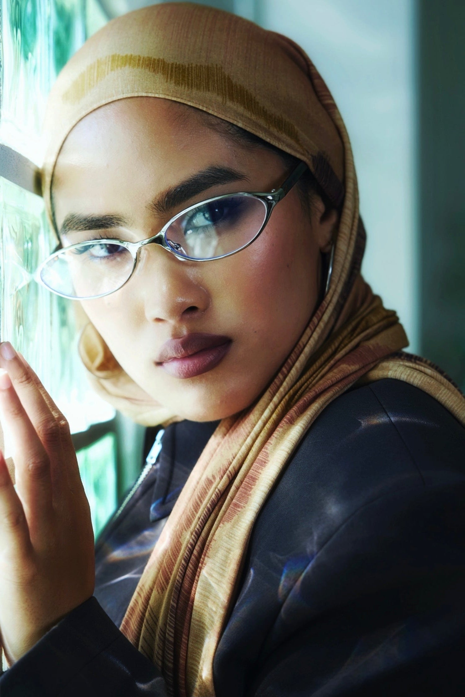 A close-up lifestyle shot of a woman wearing a beige and brown patterned hijab, silver-framed eyeglasses, and a black leather jacket, resting her head on a reflective surface.