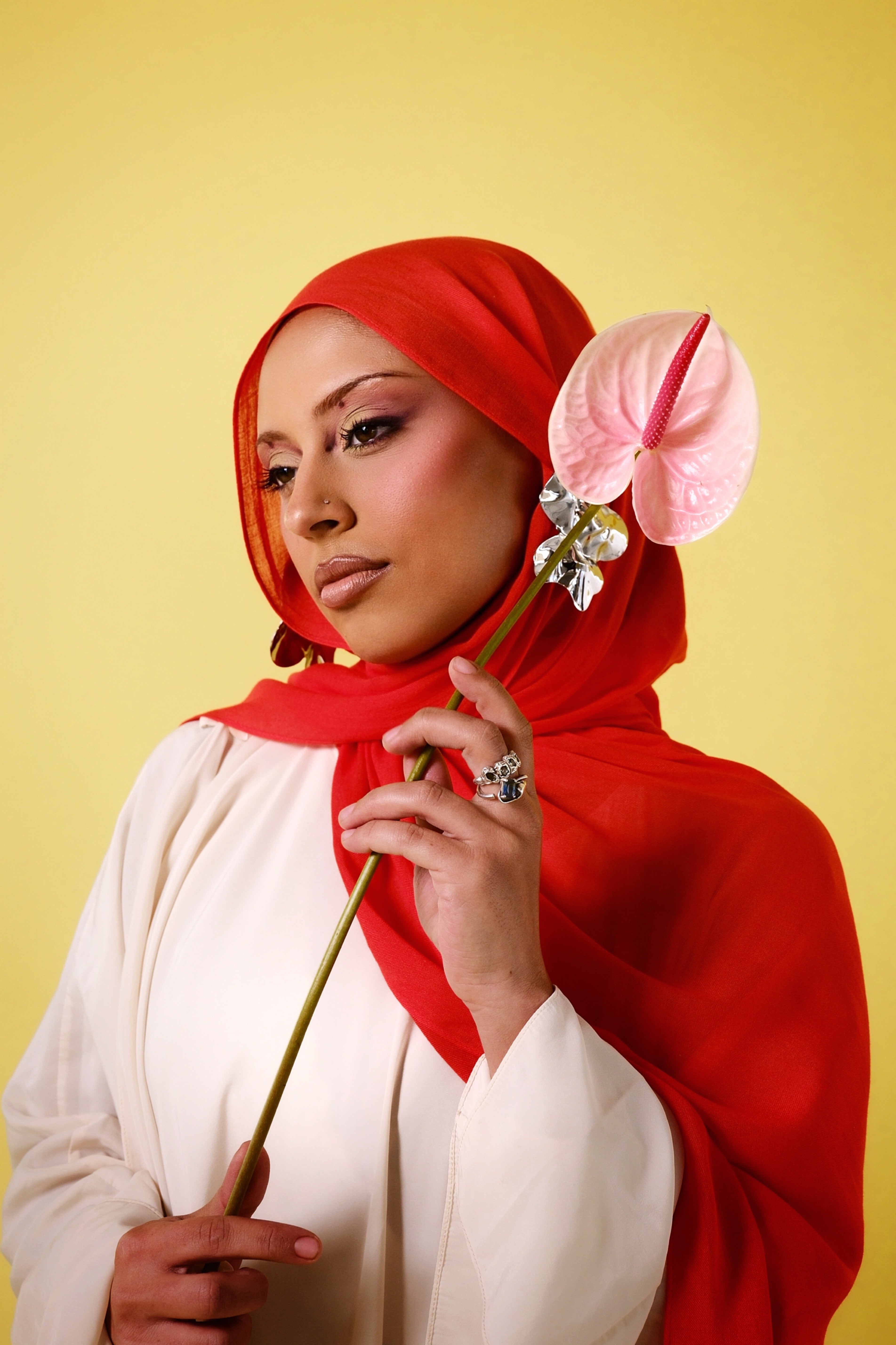 Woman wearing a red hijab holding a pink flower against a yellow background