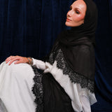 A woman wearing a black hijab and a white dress with black lace trim, posed against a dark blue velvet curtain backdrop.