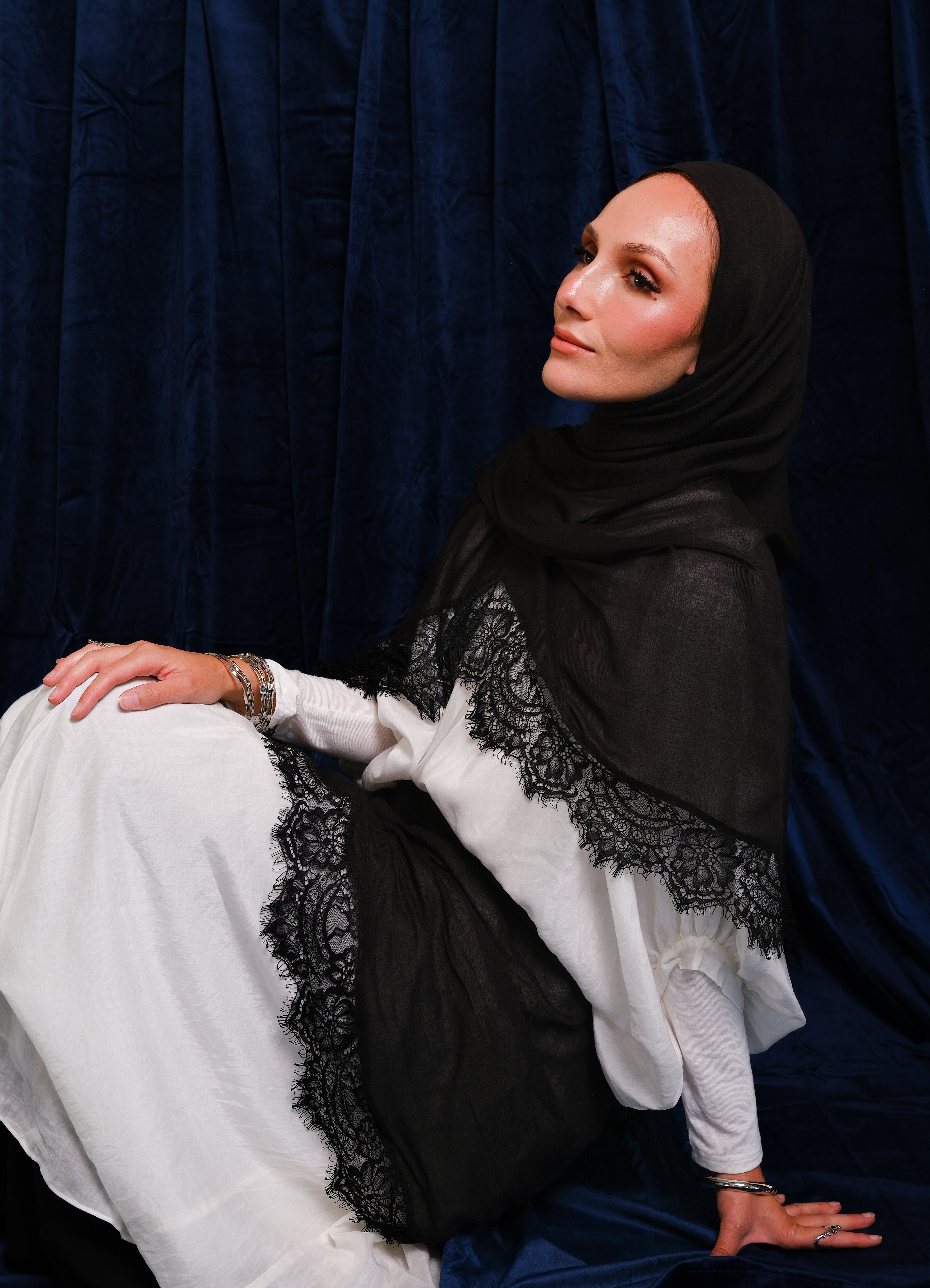 A woman wearing a black hijab and a white dress with black lace trim, posed against a dark blue velvet curtain backdrop.