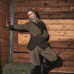 A woman wearing a vintage-style olive green jacket, brown trousers, and a beige hijab poses inside a rustic wooden barn with hay on the floor.