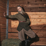 A woman wearing a vintage-style olive green jacket, brown trousers, and a beige hijab poses inside a rustic wooden barn with hay on the floor.