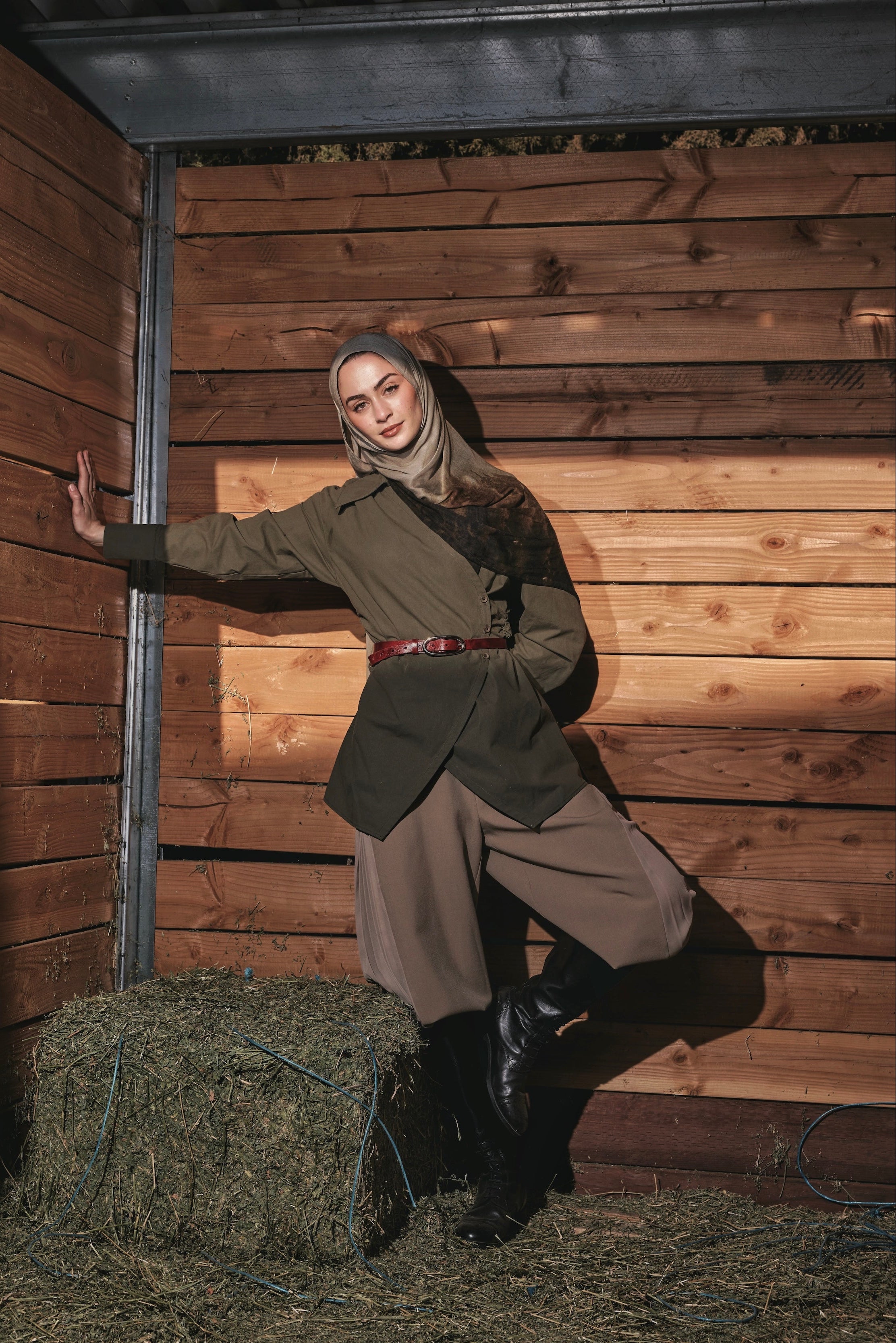 A woman wearing a vintage-style olive green jacket, brown trousers, and a beige hijab poses inside a rustic wooden barn with hay on the floor.