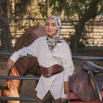 A woman wearing a white long-sleeve shirt, brown wide-leg pants, a brown leather belt, and a black-and-white patterned hijab leans against a fence in an outdoor ranch setting with a horse and a brown leather saddle nearby.