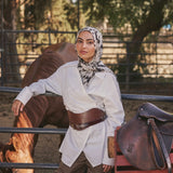 A woman wearing a white long-sleeve shirt, brown wide-leg pants, a brown leather belt, and a black-and-white patterned hijab leans against a fence in an outdoor ranch setting with a horse and a brown leather saddle nearby.