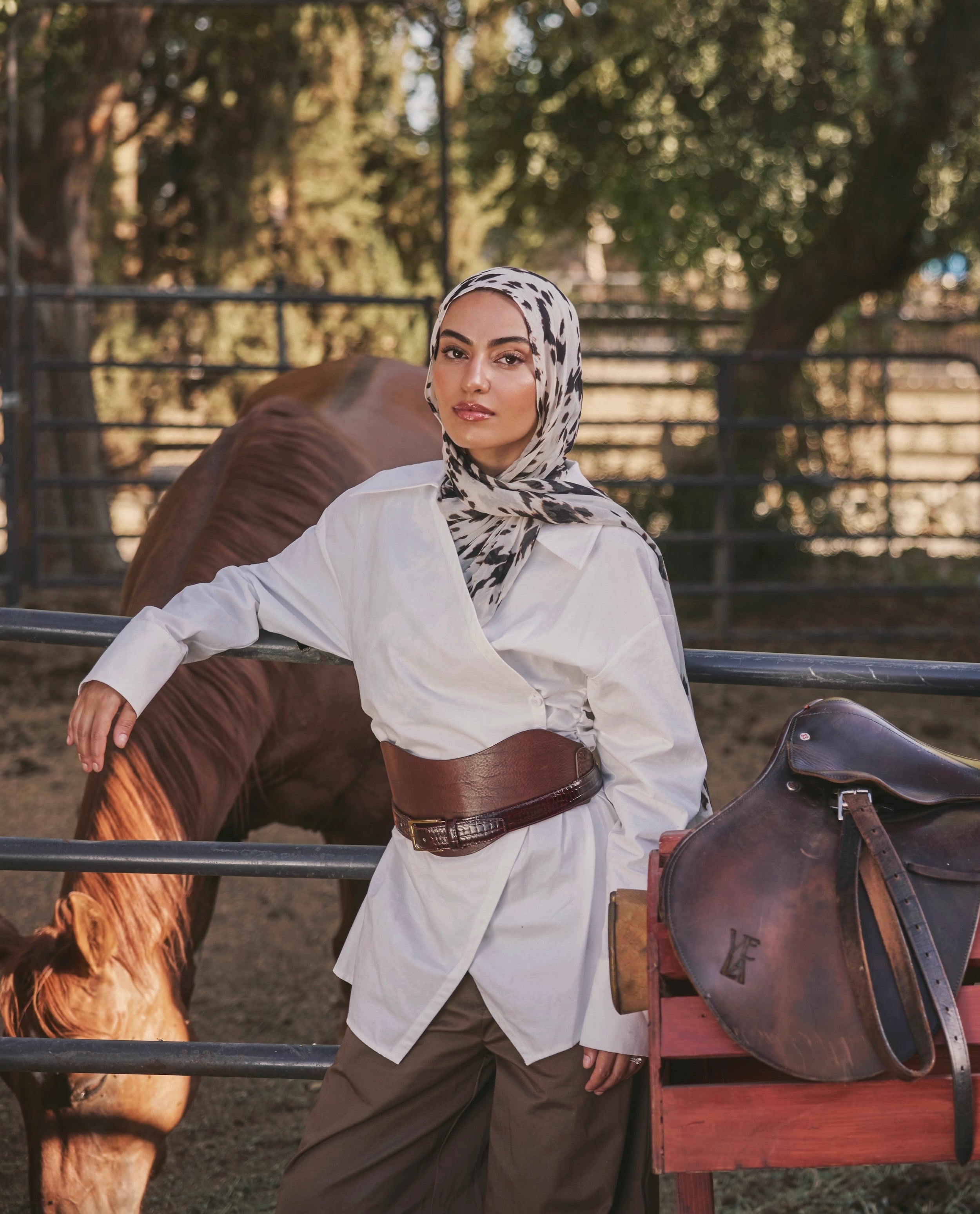 A woman wearing a white long-sleeve shirt, brown wide-leg pants, a brown leather belt, and a black-and-white patterned hijab leans against a fence in an outdoor ranch setting with a horse and a brown leather saddle nearby.