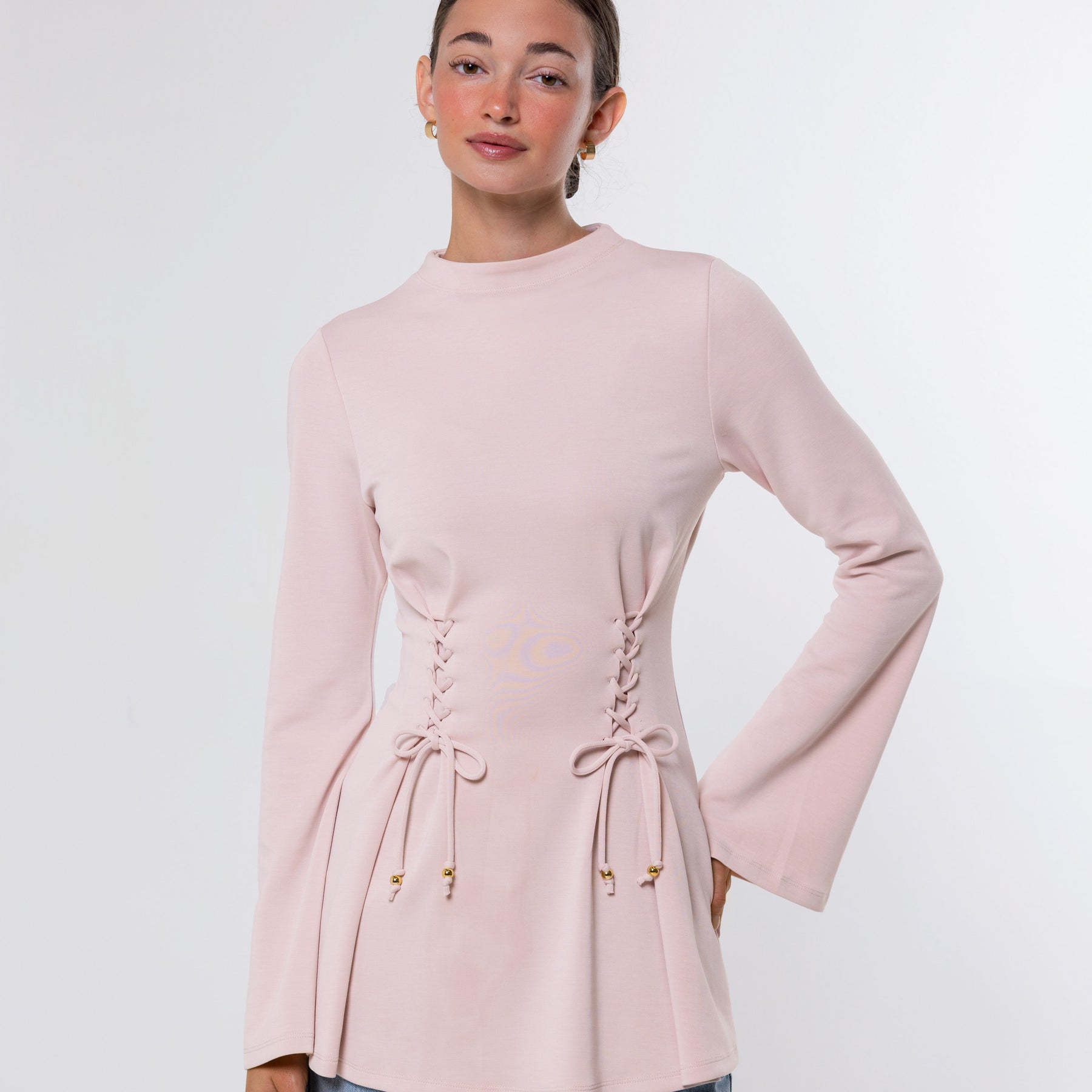 A woman modeling a light pink long-sleeve top with lace-up details at the waist and flared cuffs, paired with light blue jeans, against a plain light gray studio background.