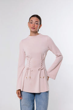 A woman modeling a light pink long-sleeve top with lace-up details at the waist and flared cuffs, paired with light blue jeans, against a plain light gray studio background.