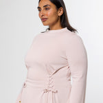 A woman wearing a pale blush pink long-sleeve dress with a lace-up corset-style waist and drawstring ties, photographed against a clean light gray studio background.