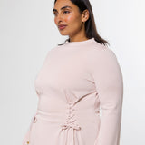 A woman wearing a pale blush pink long-sleeve dress with a lace-up corset-style waist and drawstring ties, photographed against a clean light gray studio background.