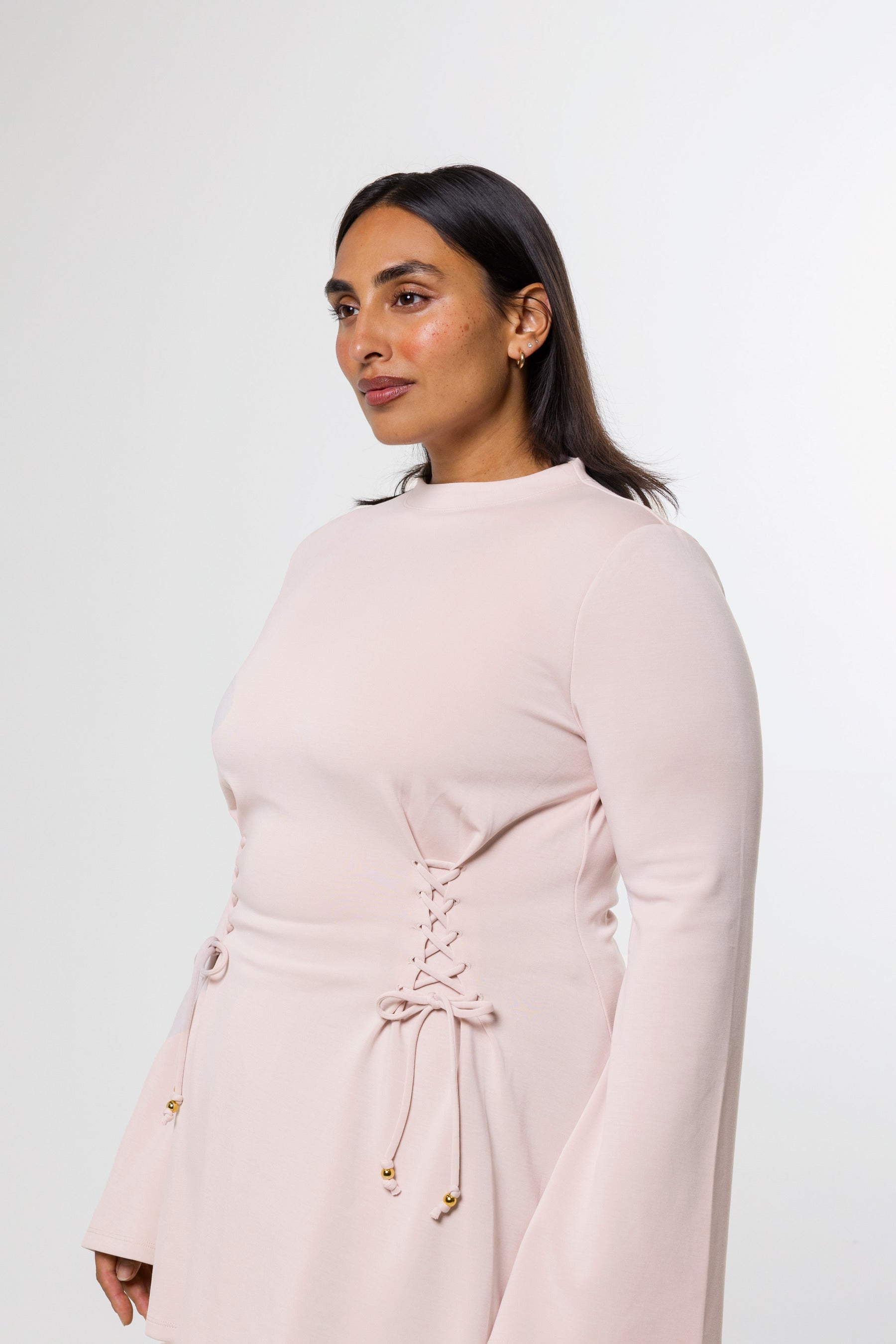 A woman wearing a pale blush pink long-sleeve dress with a lace-up corset-style waist and drawstring ties, photographed against a clean light gray studio background.