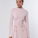 A woman modeling a light pink long-sleeve knit top with lace-up front detailing and flared cuffs, paired with light blue jeans, against a plain light gray studio background.