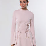 A woman modeling a light pink long-sleeve knit top with lace-up front detailing and flared cuffs, paired with light blue jeans, against a plain light gray studio background.