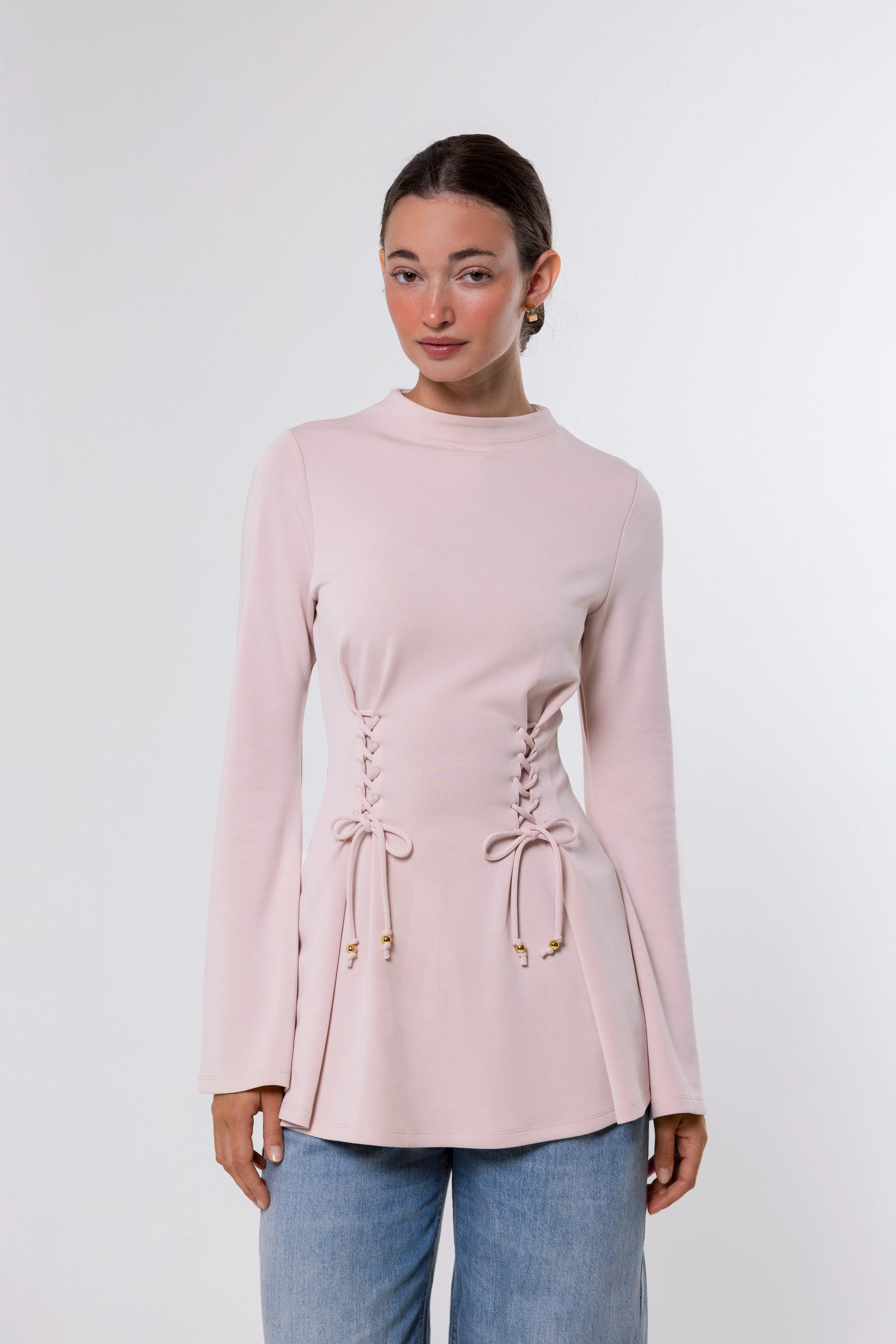A woman modeling a light pink long-sleeve knit top with lace-up front detailing and flared cuffs, paired with light blue jeans, against a plain light gray studio background.