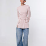 A woman modeling a light pink long-sleeve knit top with side lace-up detailing and drawstring ties, paired with wide-leg blue jeans and dark brown shoes, against a plain white studio background.