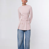 A woman modeling a light pink long-sleeve knit top with side lace-up detailing and drawstring ties, paired with wide-leg blue jeans and dark brown shoes, against a plain white studio background.