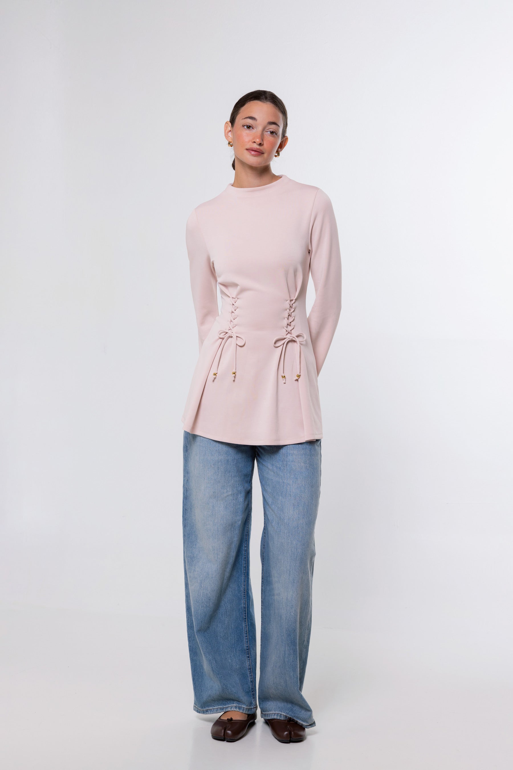 A woman modeling a light pink long-sleeve knit top with side lace-up detailing and drawstring ties, paired with wide-leg blue jeans and dark brown shoes, against a plain white studio background.