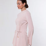 A woman shown in side profile wearing a light pink long-sleeve top with a lace-up detail at the waist and blue jeans, photographed against a plain light gray studio background.