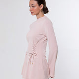 A woman shown in side profile wearing a light pink long-sleeve top with a lace-up detail at the waist and blue jeans, photographed against a plain light gray studio background.
