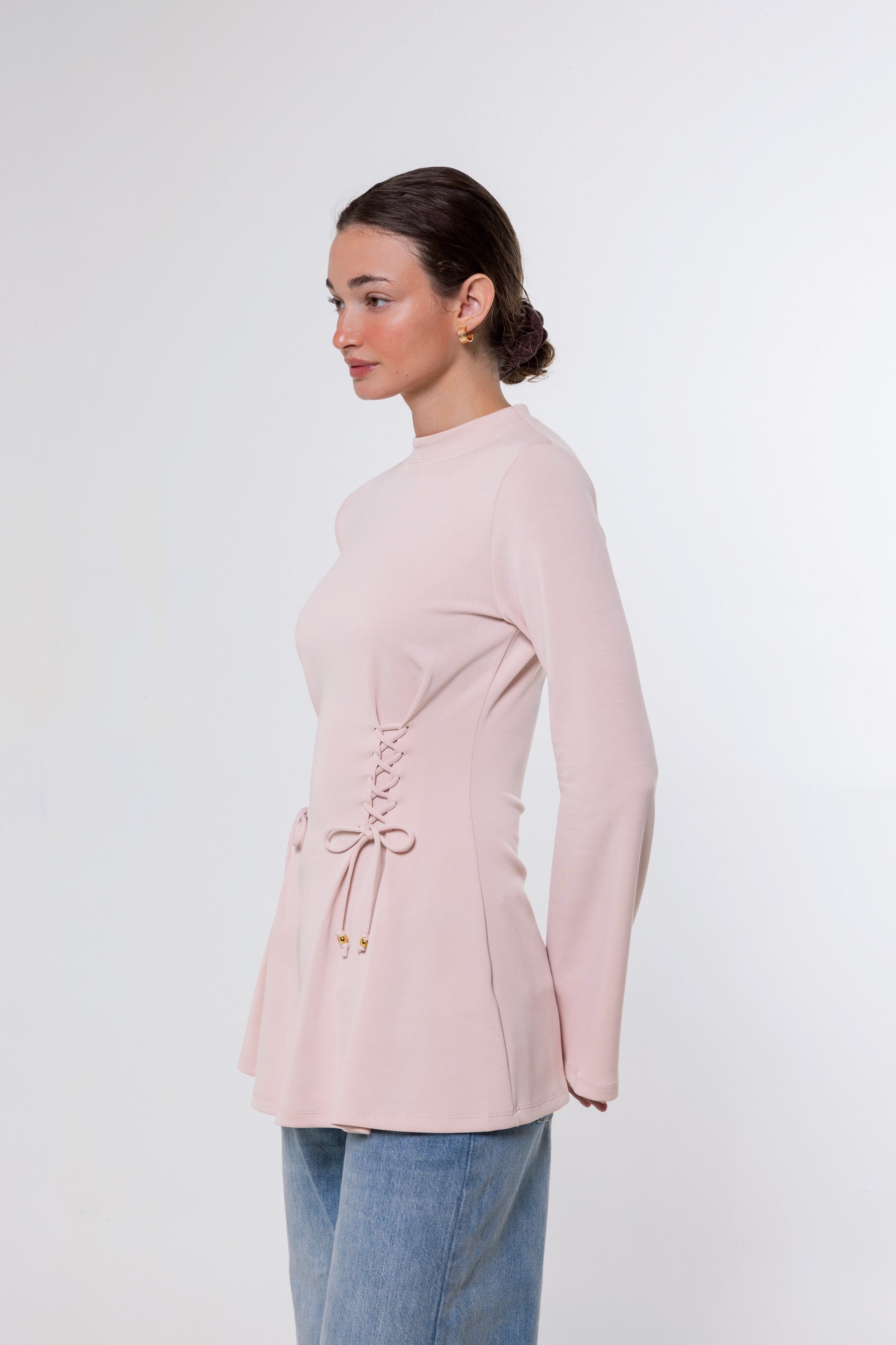 A woman shown in side profile wearing a light pink long-sleeve top with a lace-up detail at the waist and blue jeans, photographed against a plain light gray studio background.