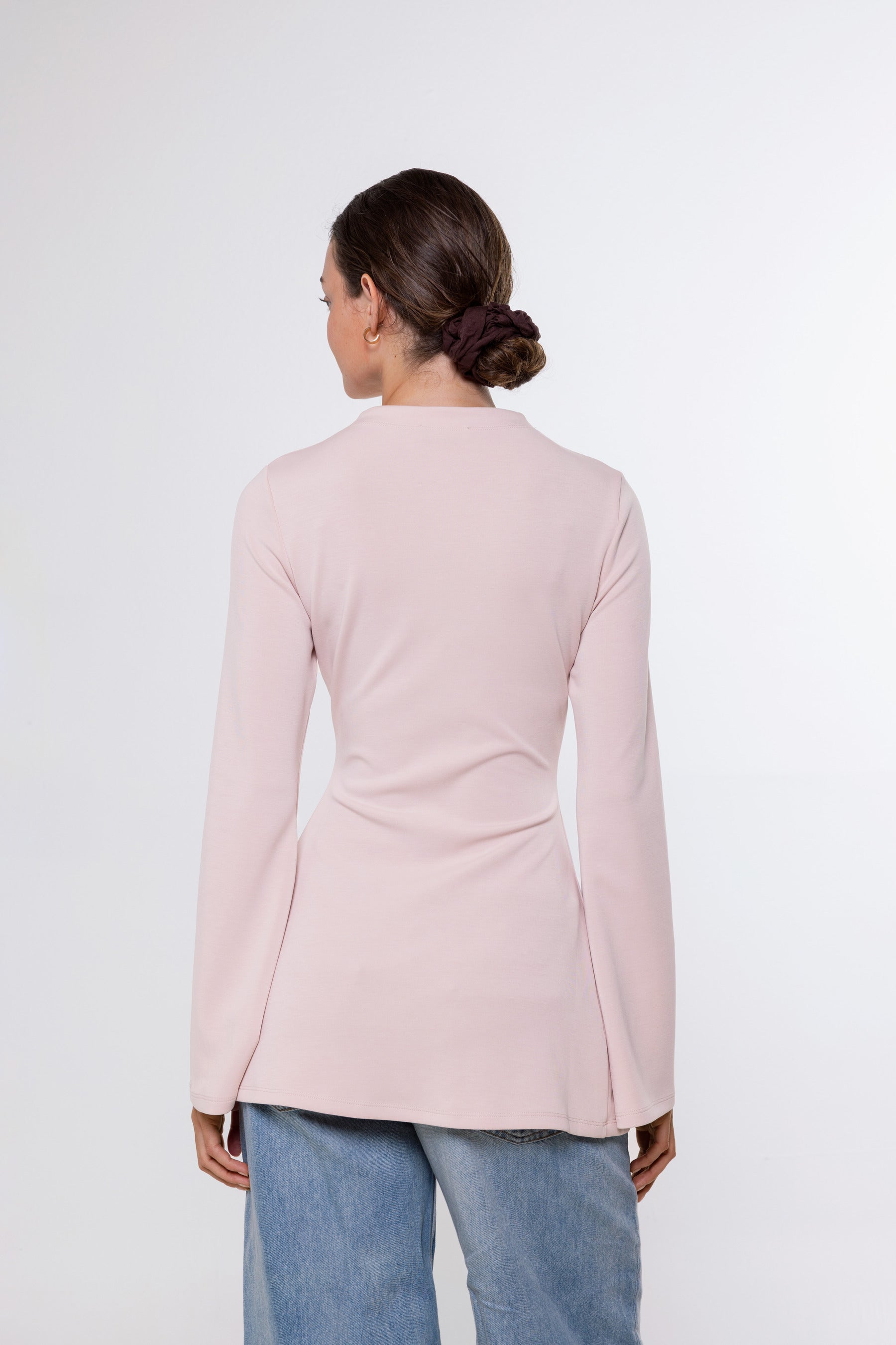 A woman viewed from the back wearing a light pink long-sleeve top and blue jeans against a plain white studio background.