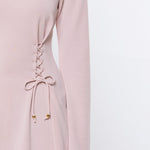 A close-up of a model wearing a light pink long-sleeve top with a lace-up detail on the side, paired with blue jeans against a plain white background.