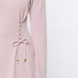A close-up of a model wearing a light pink long-sleeve top with a lace-up detail on the side, paired with blue jeans against a plain white background.