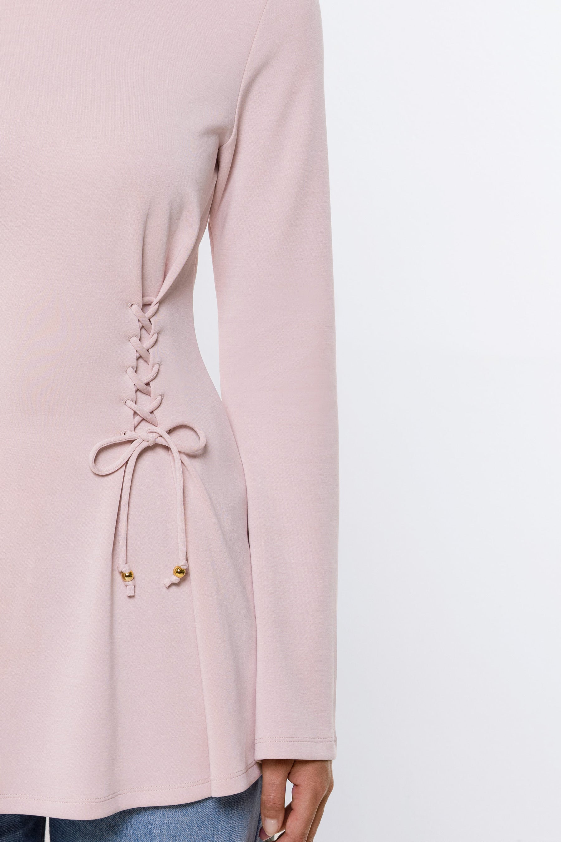 A close-up of a model wearing a light pink long-sleeve top with a lace-up detail on the side, paired with blue jeans against a plain white background.