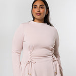 A woman modeling a light pink long-sleeve top with a gathered waist and lace-up front detail, set against a plain light gray studio background.