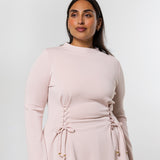 A woman modeling a light pink long-sleeve top with a gathered waist and lace-up front detail, set against a plain light gray studio background.