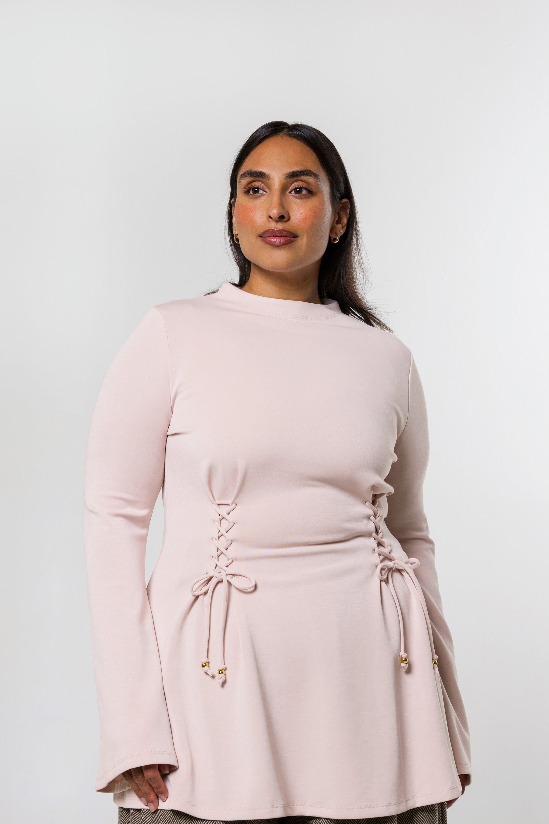 A woman modeling a light pink long-sleeve top with a gathered waist and lace-up front detail, set against a plain light gray studio background.