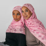 Two women wearing pink floral hijabs against a plain background