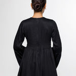 A woman viewed from the back wearing a black long-sleeve peplum-style top with textured vertical pleating on the back, paired with light blue jeans, against a plain light gray studio background.