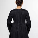 A woman viewed from the back wearing a black long-sleeve peplum-style top with textured vertical pleating on the back, paired with light blue jeans, against a plain light gray studio background.