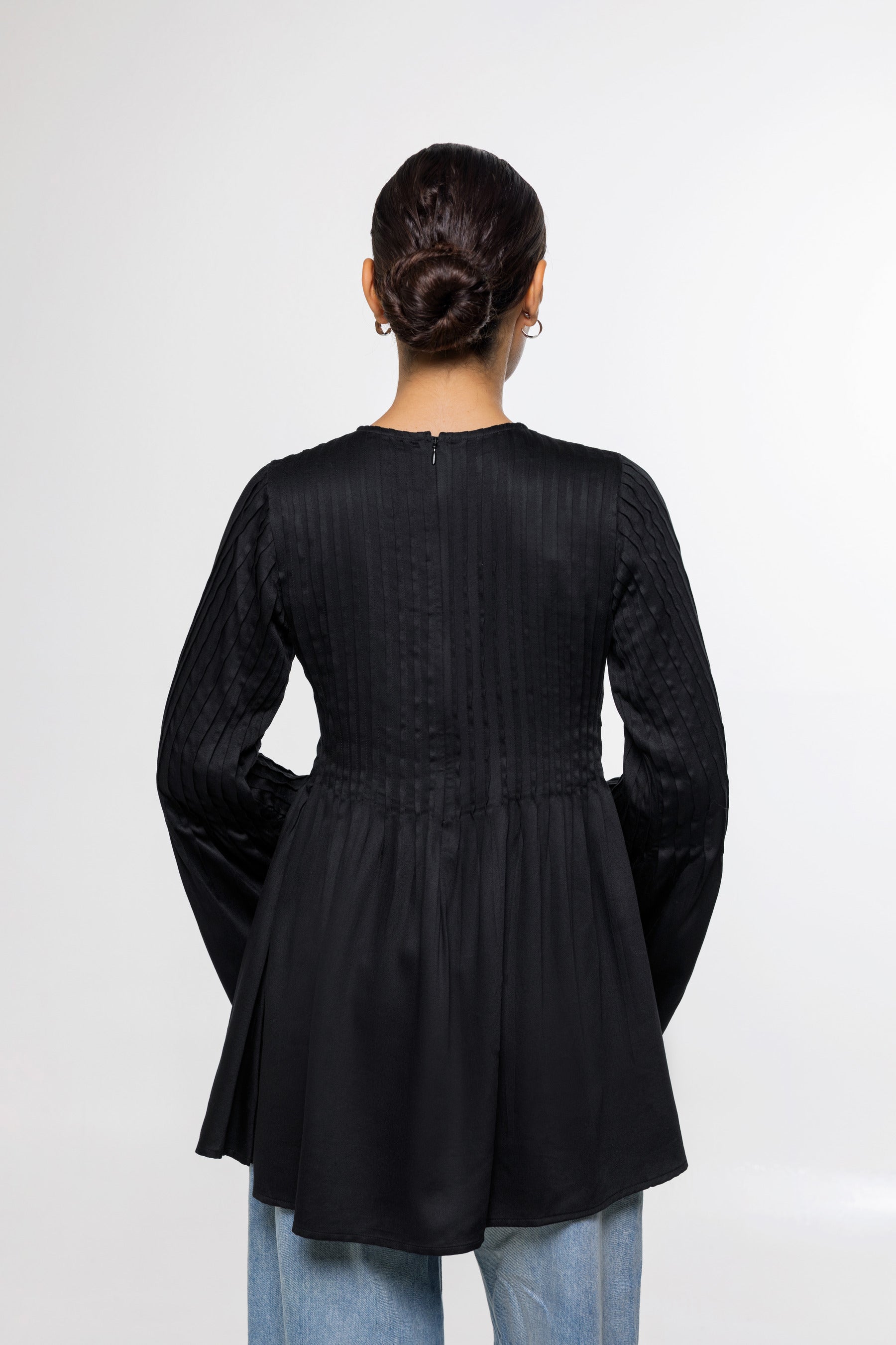 A woman viewed from the back wearing a black long-sleeve peplum-style top with textured vertical pleating on the back, paired with light blue jeans, against a plain light gray studio background.