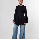 A woman modeling a black long-sleeve top with a gathered waist and flared bell sleeves, paired with wide-leg blue jeans and black pointed shoes, against a light gray studio background.