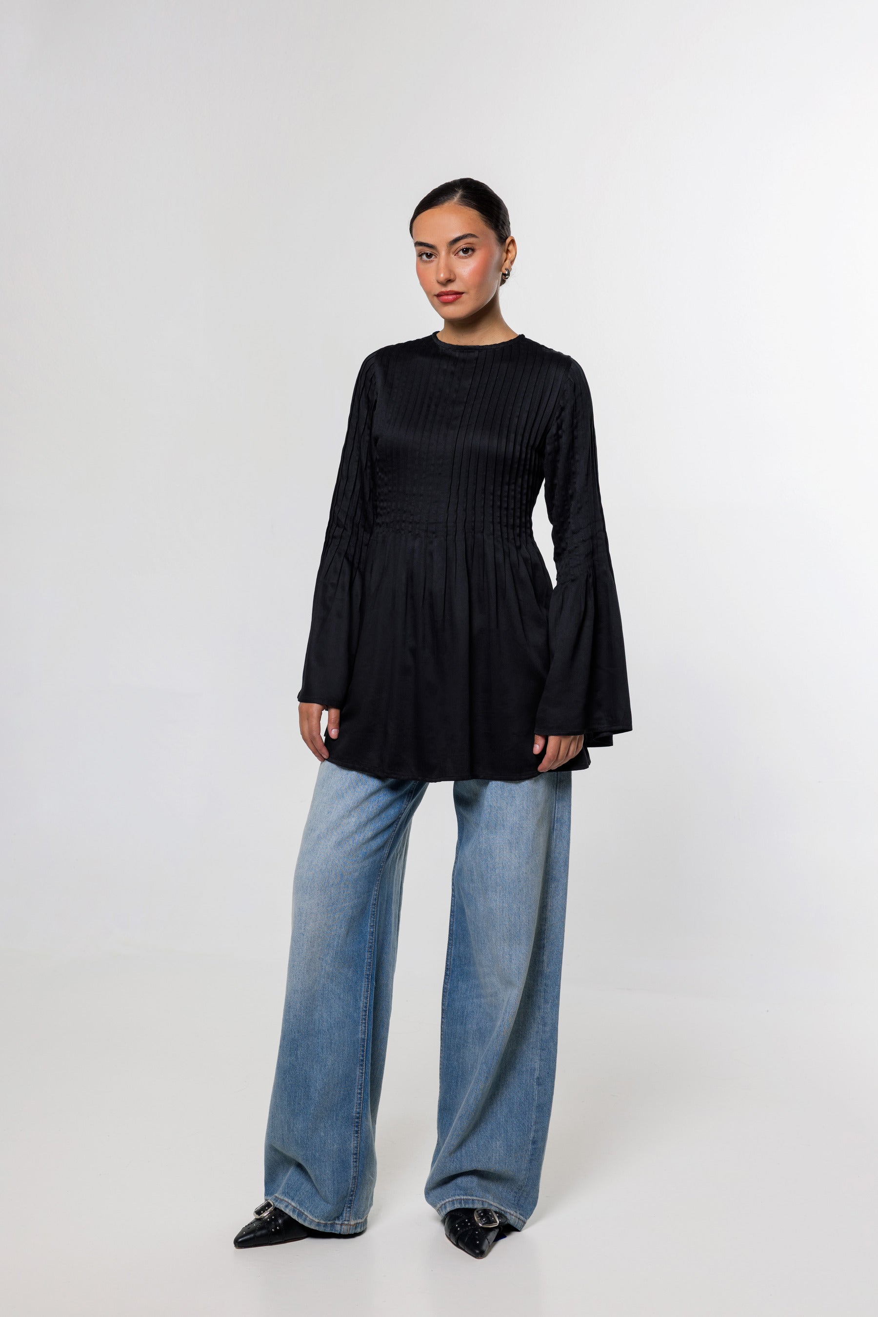 A woman modeling a black long-sleeve top with a gathered waist and flared bell sleeves, paired with wide-leg blue jeans and black pointed shoes, against a light gray studio background.