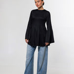 A woman modeling a black long-sleeve top with a gathered peplum waist and flared cuffs, paired with wide-leg blue jeans and black shoes, against a plain light gray studio background.