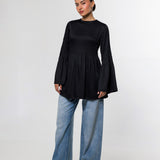 A woman modeling a black long-sleeve top with a gathered peplum waist and flared cuffs, paired with wide-leg blue jeans and black shoes, against a plain light gray studio background.