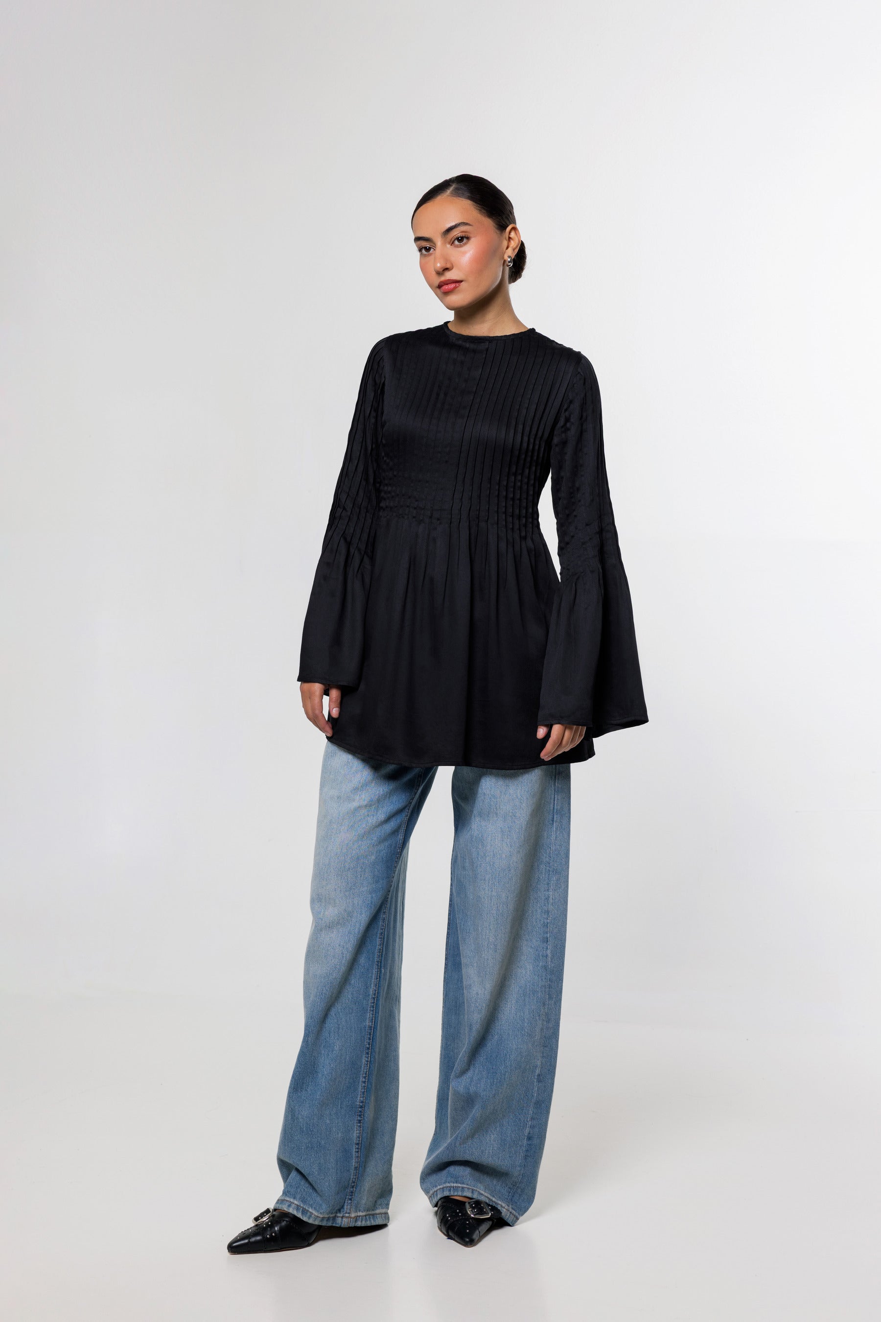 A woman modeling a black long-sleeve top with a gathered peplum waist and flared cuffs, paired with wide-leg blue jeans and black shoes, against a plain light gray studio background.