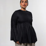 A woman modeling a black long-sleeve top with a textured vertical pleated bodice and flared sleeves, paired with light beige trousers, against a plain light gray studio background.