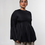A woman modeling a black long-sleeve top with a textured vertical pleated bodice and flared sleeves, paired with light beige trousers, against a plain light gray studio background.