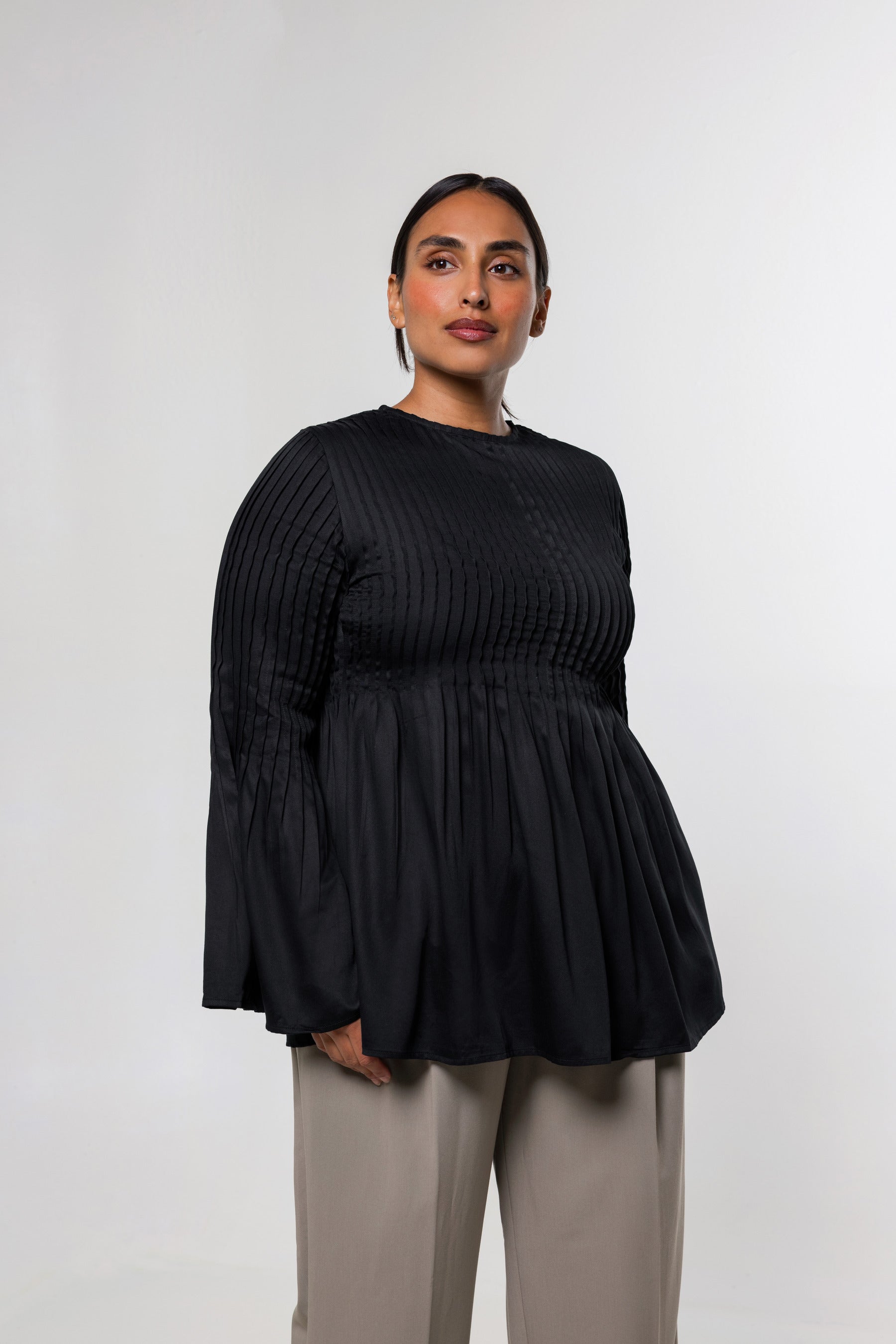 A woman modeling a black long-sleeve top with a textured vertical pleated bodice and flared sleeves, paired with light beige trousers, against a plain light gray studio background.