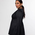 A woman modeling a black pleated long-sleeve top with a flared peplum hem, paired with beige trousers, photographed against a plain light gray studio background.
