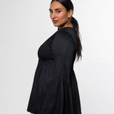 A woman modeling a black pleated long-sleeve top with a flared peplum hem, paired with beige trousers, photographed against a plain light gray studio background.