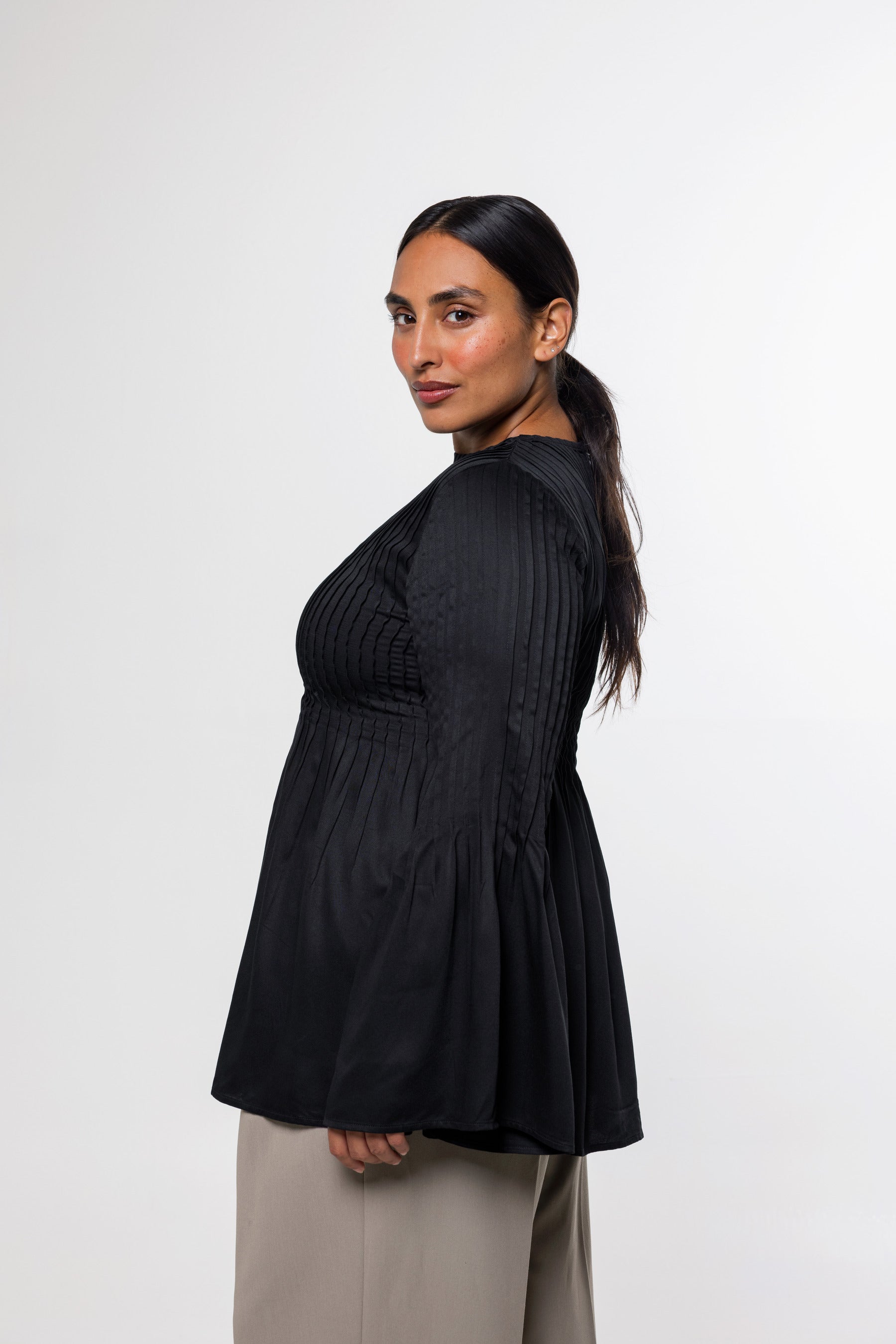 A woman modeling a black pleated long-sleeve top with a flared peplum hem, paired with beige trousers, photographed against a plain light gray studio background.